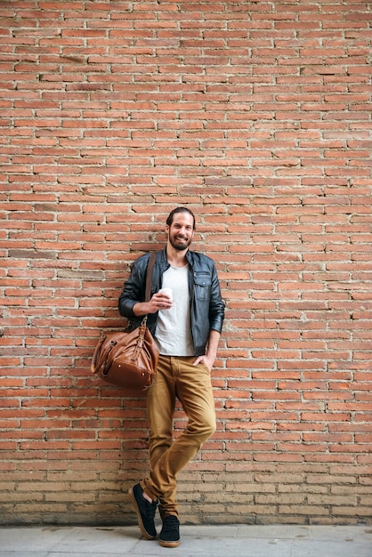 Retrato de corpo inteiro de um homem europeu sorridente com cabelo amarrado, olhando para você, em pé sobre um muro de tijolos na rua da cidade e bebendo café para viagem