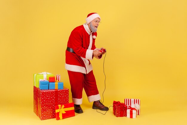 Retrato de corpo inteiro de um homem idoso com barba grisalha, vestindo fantasia de Papai Noel, jogando videogame na véspera de ano novo com expressão concentrada. Estúdio interno, tiro isolado em fundo amarelo.