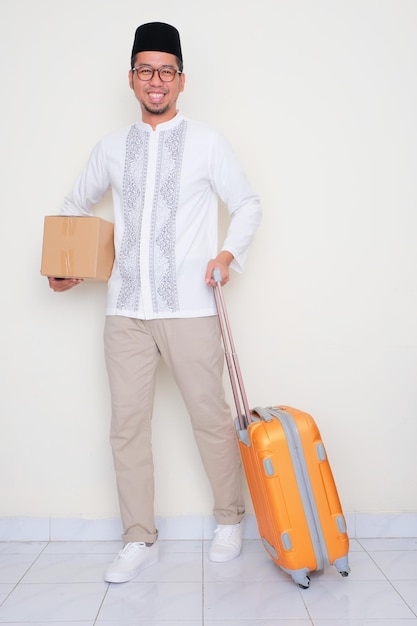 Retrato de corpo inteiro de um homem muçulmano sorrindo enquanto traz bagagem e carrega uma caixa de papelão