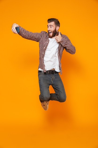 Retrato de corpo inteiro de um jovem alegre, vestindo roupas casuais, pulando e tirando uma selfie