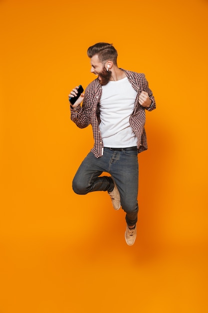 Retrato de corpo inteiro de um jovem alegre, vestindo roupas casuais, pulando ouvindo música com fones de ouvido segurando um telefone celular