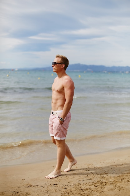 Retrato de corpo inteiro de um jovem bonito em calções de praia, caminhando na praia. Moda de viagem. Estilo de vida esportivo