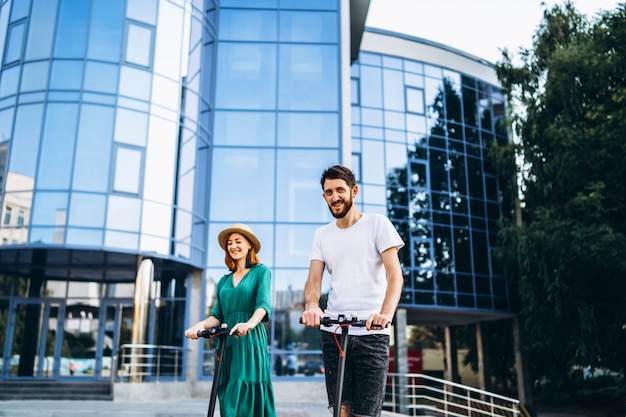 Retrato de corpo inteiro de um jovem casal romântico com scooters elétricos, andando na cidade.