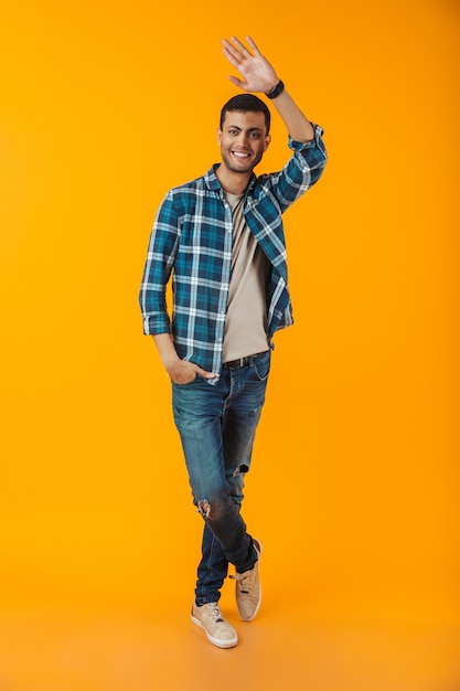 Retrato de corpo inteiro de um jovem feliz vestindo uma camisa xadrez isolada na parede laranja, acenando com a mão