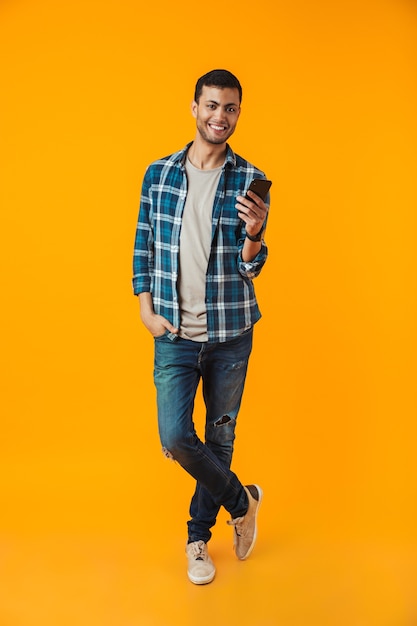 Retrato de corpo inteiro de um jovem feliz vestindo uma camisa xadrez isolada sobre um fundo laranja, usando um telefone celular