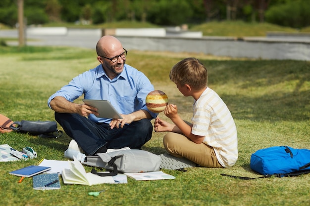 Retrato de corpo inteiro de um professor careca sorridente falando com um adolescente segurando o planeta modelo enquanto desfruta de uma aula de astronomia ao ar livre sob a luz do sol, copie o espaço