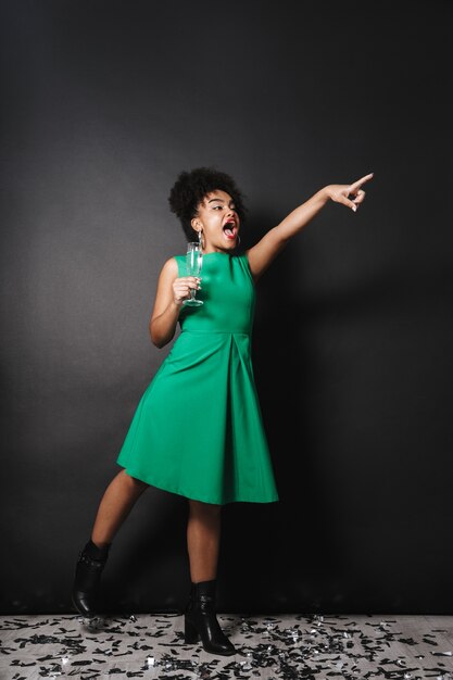 Retrato de corpo inteiro de uma alegre mulher afro-americana usando um vestido em pé sobre uma parede preta