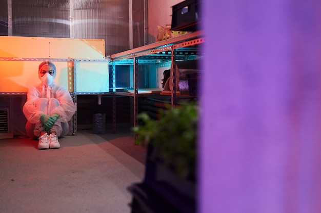 Retrato de corpo inteiro de uma cientista usando equipamento de proteção completo, sentada no chão de um laboratório biológico iluminado por luz ultravioleta, copie o espaço