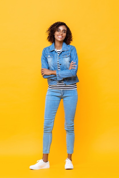 Retrato de corpo inteiro de uma jovem afro-americana sorridente em pé com os braços cruzados no fundo do estúdio amarelo