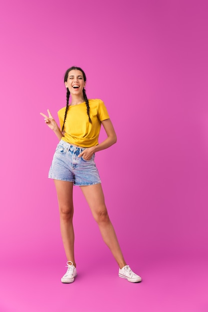 Retrato de corpo inteiro de uma jovem alegre, vestindo shorts jeans, piscando e gesticulando para o sinal da paz isolado na parede rosa