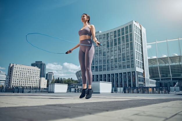 Retrato de corpo inteiro de uma jovem em forma em roupas esportivas da moda, se exercitando com uma corda de pular