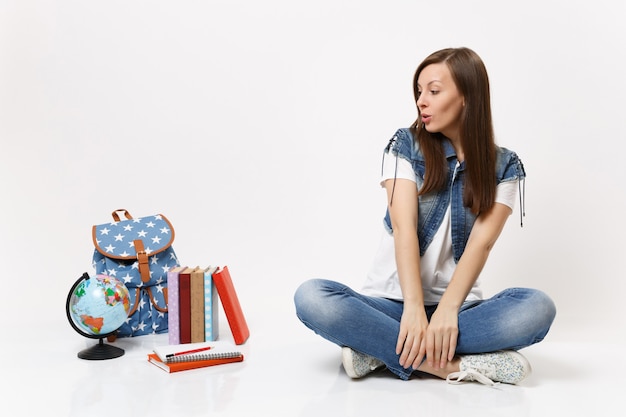 Retrato de corpo inteiro de uma jovem estudante casual em roupas jeans, sentada olhando no globo, mochila, livros escolares isolados