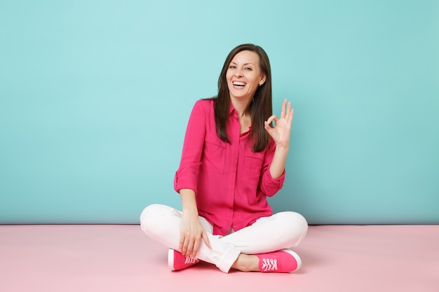 Retrato de corpo inteiro de uma jovem sorridente com blusa de camisa rosa e calça branca sentada no chão