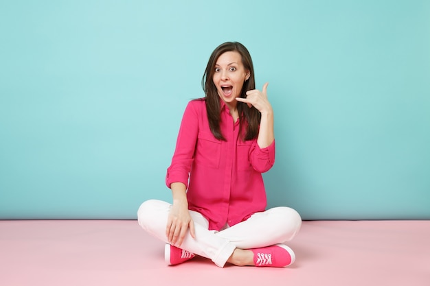 Retrato de corpo inteiro de uma jovem sorridente com blusa de camisa rosa e calça branca sentada no chão