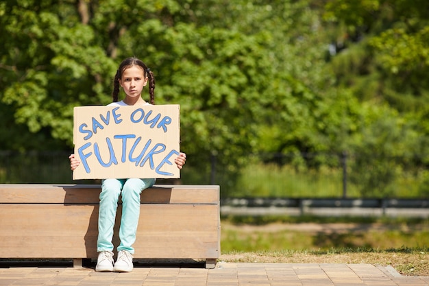 Retrato de corpo inteiro de uma linda garota segurando uma placa com a escrita SALVAR FUTURO enquanto protesta pela natureza e pela economia ao ar livre, copie o espaço