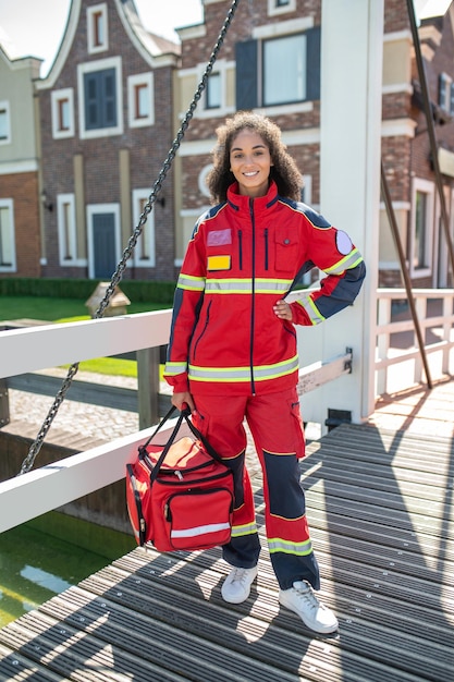 Retrato de corpo inteiro de uma médica sorridente com uma bolsa EMS na mão, olhando para o futuro