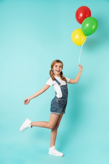 Retrato de corpo inteiro de uma menina alegre isolada sobre uma parede azul, segurando um monte de balões de ar coloridos, posando