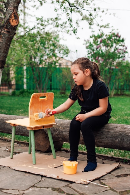 Retrato de corpo inteiro de uma menina em roupas pretas ajudando os pais a pintar uma cadeira para um irmão ou irmã ...