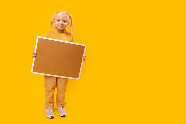 Retrato de corpo inteiro de uma menina feliz com uma placa de cortiça vazia em fundo amarelo Copie o espaço simulado