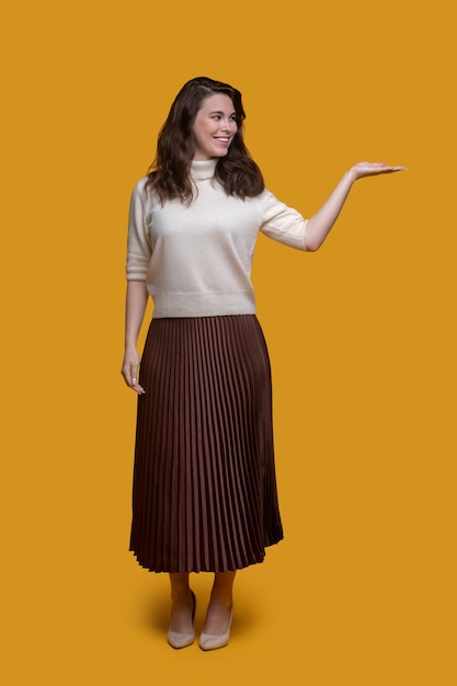 Retrato de corpo inteiro de uma mulher alegre e moderna vestida com uma longa saia plissada segurando a palma esquerda para cima