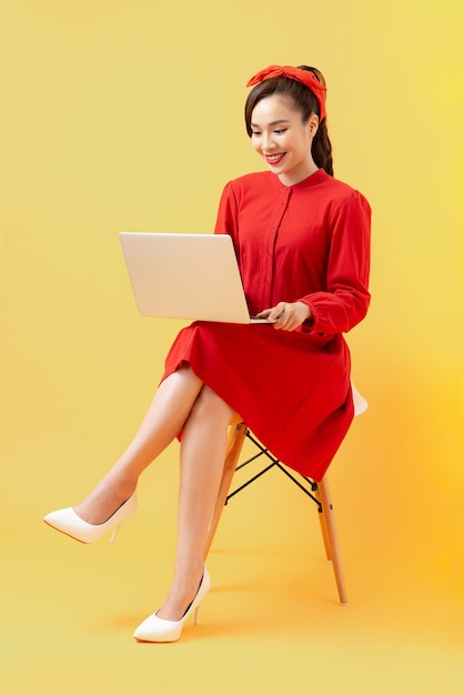 Retrato de corpo inteiro de uma mulher asiática casual feliz segurando um laptop enquanto está sentado em uma cadeira sobre um fundo laranja