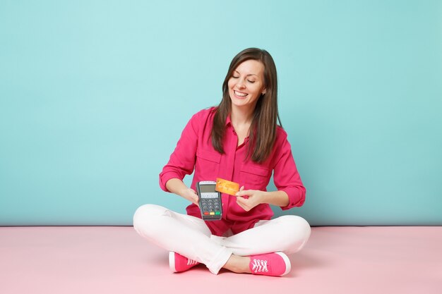 Retrato de corpo inteiro de uma mulher de camisa rosa e calça branca sentada no chão com cartão terminal