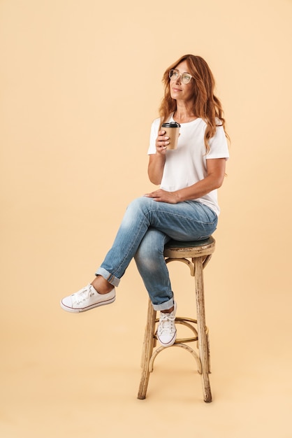 Foto retrato de corpo inteiro de uma mulher de meia-idade sorridente atraente, vestindo roupa casual, sentada em uma cadeira isolada sobre uma parede bege, segurando um café para viagem