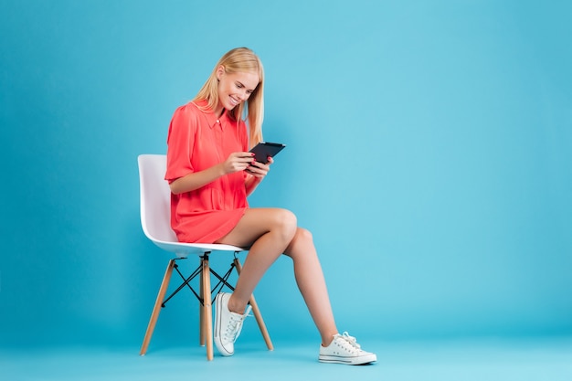 Retrato de corpo inteiro de uma mulher loira sorridente com vestido vermelho, sentada na cadeira e usando um tablet isolado na parede azul