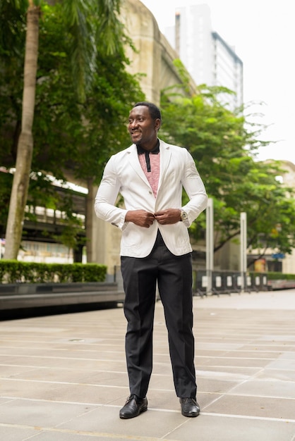 Retrato de corpo inteiro do bonito empresário africano na cidade, sorrindo e pensando