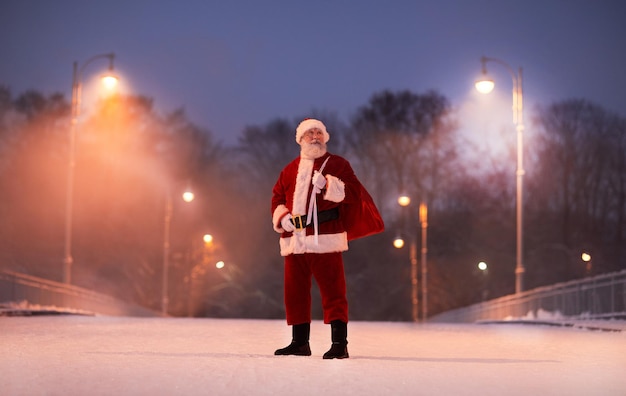 Retrato de corpo inteiro do tradicional papai noel em pé na rua da cidade à noite na véspera de natal co