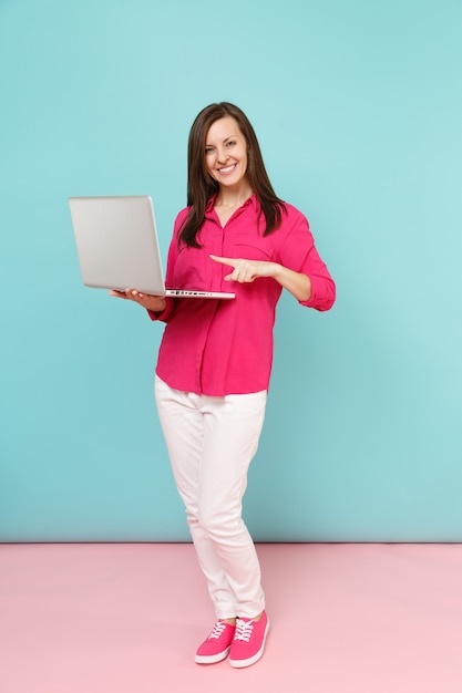 Retrato de corpo inteiro jovem com blusa de camisa rosa, calça branca, segurando o computador laptop pc isolado na parede azul pastel de rosa brilhante.