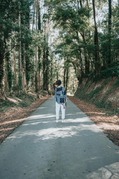 Retrato de corpo inteiro jovem por trás andando com prancha longa no meio da floresta