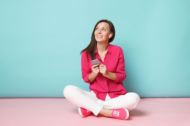 Retrato de corpo inteiro mulher com blusa de camisa rosa e calça branca sentada no chão com fones de ouvido