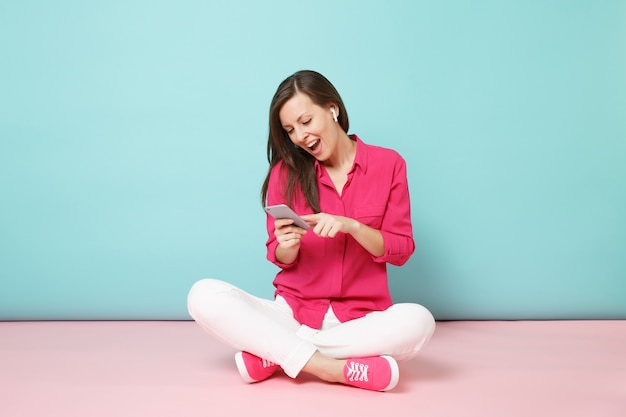 Retrato de corpo inteiro mulher com blusa de camisa rosa e calça branca sentada no chão com fones de ouvido