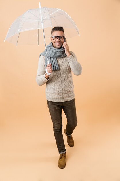 Retrato de corpo inteiro se um homem feliz, vestido com um suéter e um lenço, caminhando sob o guarda-chuva, isolado sobre uma parede bege, falando no celular