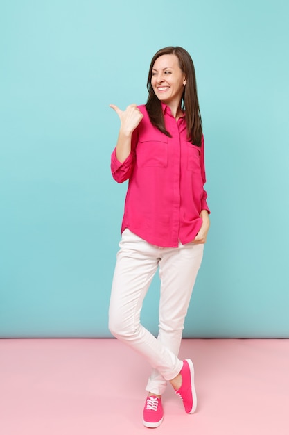 Retrato de corpo inteiro sorridente jovem bonita na blusa de camisa rosa, calça branca posando isolado na parede pastel azul rosa brilhante.