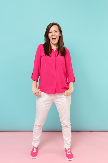 Retrato de corpo inteiro sorridente jovem bonita na blusa de camisa rosa, calça branca posando isolado na parede pastel azul rosa brilhante.