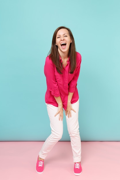 Retrato de corpo inteiro sorridente jovem bonita na blusa de camisa rosa, calça branca posando isolado na parede pastel azul rosa brilhante.