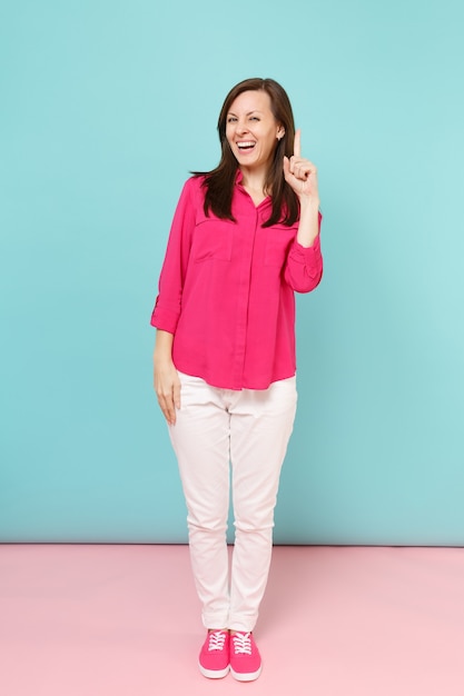 Retrato de corpo inteiro sorridente jovem bonita na blusa de camisa rosa, calça branca posando isolado na parede pastel azul rosa brilhante.