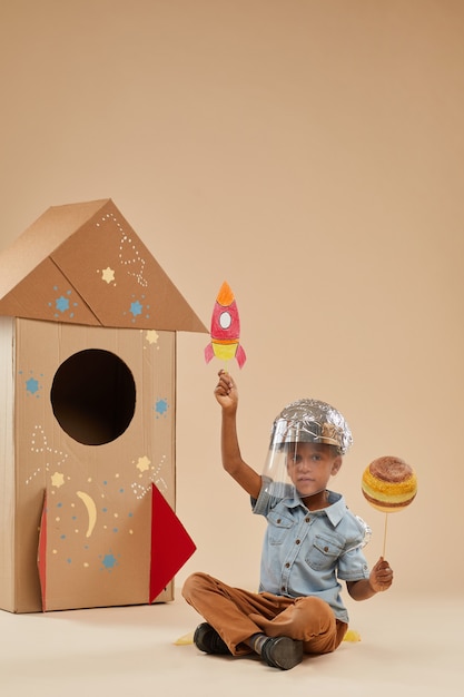 Retrato de corpo inteiro vertical de um menino afro-americano fofo brincando de astronauta em um traje espacial feito à mão, sentado no chão em um foguete de papelão