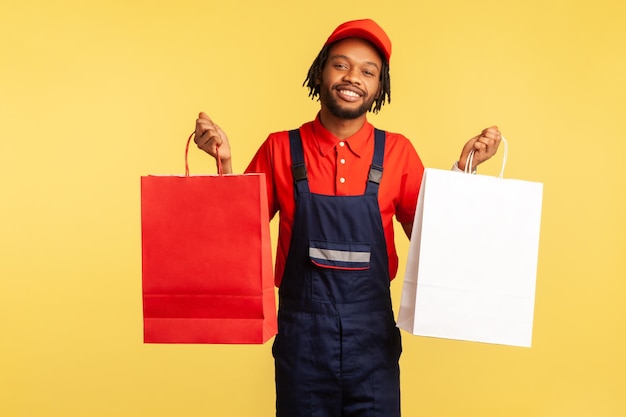 Retrato de correio otimista em uniforme segurando sacolas de compras e parece feliz entregando encomendas com mercadorias encomendadas on-line na loja de moda Estúdio interior isolado em fundo amarelo