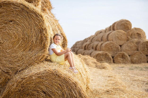 Retrato de criança do sexo feminino alegre vestindo vestido no alto palheiro, apoiando-se em outro palheiro com muitos outros palheiros no fundo se divertindo longe da cidade no campo cheio de feno dourado