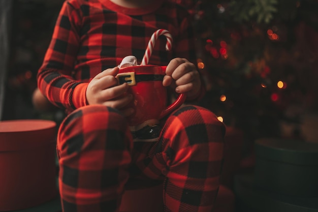 Retrato de criança feliz e sincera em pijama xadrez vermelho segura caneca de Natal com marshmallows e cana-de-doce