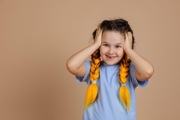 Retrato de criança feliz surpresa com as mãos tocando a cabeça com sorriso com dente perdido, olhando para a câmera com tranças kanekalon amarelas sobre fundo bege, vestindo jeans e camiseta azul