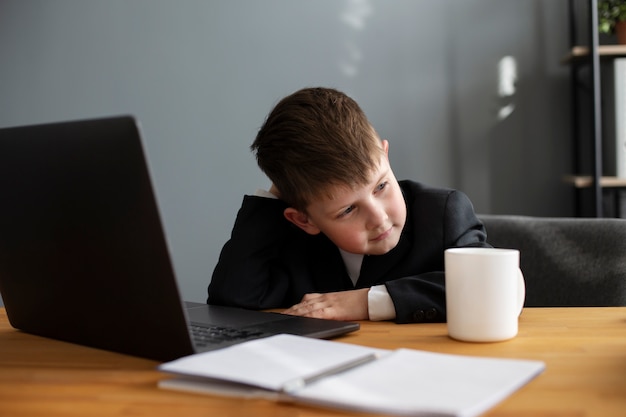 Retrato de criança fofa com terno sentado na mesa com laptop e caneca