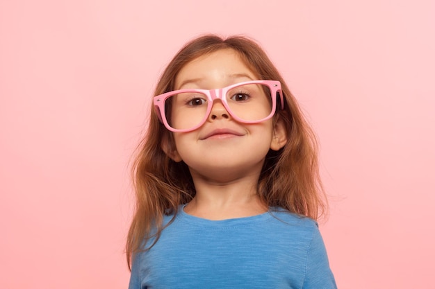 Retrato de criança gênio positivo menina inteligente usando óculos cor de rosa e olhando para a câmera com expressão feliz inteligente óculos elegante moda estúdio interior tiro isolado no fundo rosa