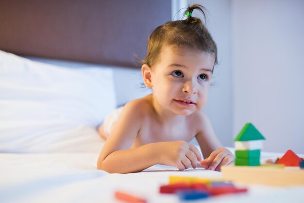 Retrato de criança linda menina deitada na cama em uma bagunça de brinquedo brincando com blocos de madeira de construção colorida na luz da lâmpada Jogo de educação para criança no quarto Criatividade do berçário