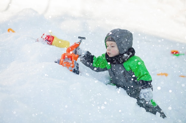 Retrato de criança pequena bonito sentado na neve e brincando com seu brinquedo trator amarelo no parque. criança brincando ao ar livre