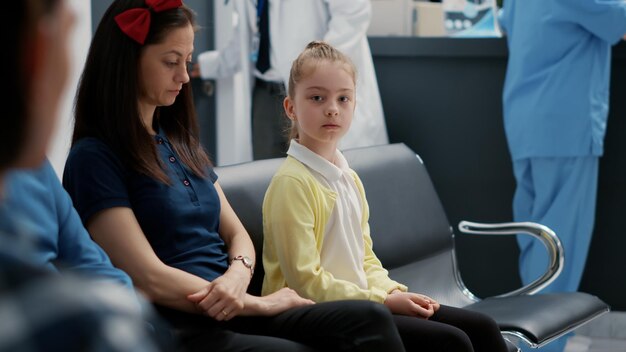 Retrato de criança sentada com a mãe na sala de espera na recepção do hospital, participando da visita anual de check-up com especialista. Menina no saguão da sala de espera no centro de saúde.