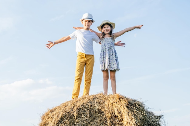 Retrato de crianças menino e menina espalhando e agitando os braços sorrindo no palheiro no campo usando chapéus de palha